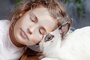 Little girl playing with real rabbit. Child and white bunny on Easter on flower background. Kid kiss pet. Fun and friendship for