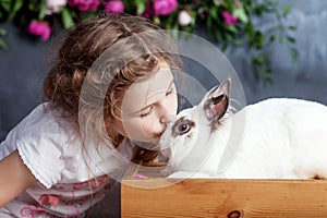 Little girl playing with real rabbit. Child and white bunny on Easter on flower background. Kid kiss pet. Fun and friendship for