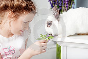 Little girl playing with real rabbit. Child and white bunny on Easter on flower background. Toddler kid feeding pet animal. Kids