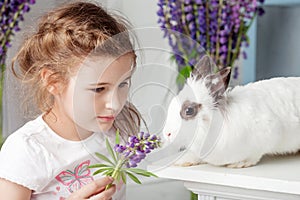 Little girl playing with real rabbit. Child and white bunny on Easter on flower background. Toddler kid feeding pet animal. Kids