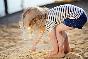 Little girl playing with sand during walk by the lake at summer day. Baby draw finger on sand