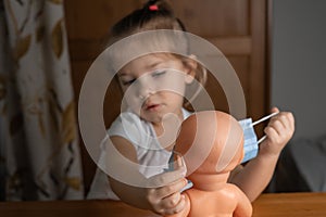 a little girl plays with a baby doll at home, puts a medical mask on a doll. focus on the doll, child in blur