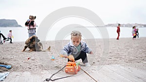 Little girl plays in the sand on the beach. Two-year-old child in a denim jacket playing with plastic toys on a sandy