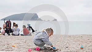 Little girl plays in the sand on the beach. Two-year-old child in a denim jacket playing with plastic toys on a sandy