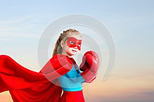 Little girl plays superhero on the background of sunset sky.