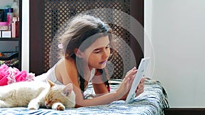 Little girl plays in the tablet in online games lies on the bed next to the cat pet. Girl and cat play tablet social
