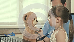 Little girl plays with a teddy bear toy listens to him with a stethoscope. toddler patient sits on lap of pediatrician