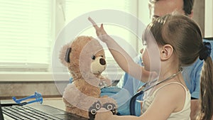 Little girl plays with a teddy bear toy listens to him with a stethoscope. toddler patient sits on lap of pediatrician