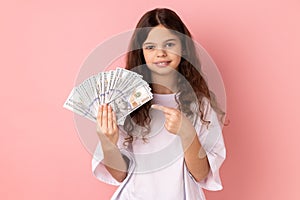 Little girl pointing at big fan of dollars banknotes with finger, rejoices of winning lottery.