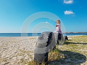 A little girl points her hand to the side while sitting near the sea on a tire buried in the ground until half