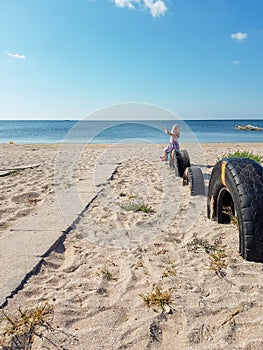 A little girl points her hand to the side while sitting near the sea on a tire buried in the ground until half