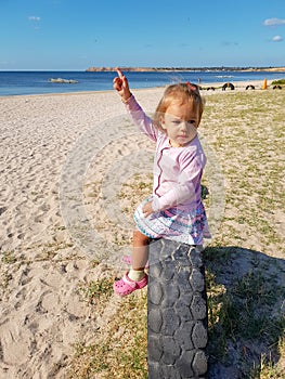 A little girl points her hand to the sky while sitting near the sea on a tire buried in the ground until half
