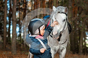 Little girl portrait, stands next to a white pony close-up on the background of nature. Jockey, epodrome, horseback riding