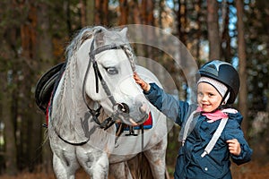 Little girl portrait, stands next to a white pony close-up on the background of nature. Jockey, epodrome, horseback riding