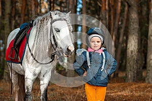 Little girl portrait, stands next to a white pony close-up on the background of nature. Jockey, epodrome, horseback riding