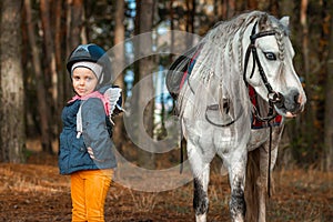 Little girl portrait, stands next to a white pony close-up on the background of nature. Jockey, epodrome, horseback riding