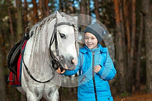 Little girl portrait, stands next to a white pony close-up on the background of nature. Jockey, epodrome, horseback riding