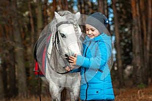 Little girl portrait, stands next to a white pony close-up on the background of nature. Jockey, epodrome, horseback riding