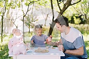 Little girl pours tea from a toy teapot into a toy cup in her daddy hand at a tea party in the garden