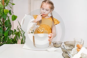 little girl prepares dough or cream in a submersible mixer and licks his fingers