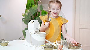 The little girl prepares dough or cream in a submersible mixer, licks the spoon