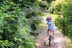 Little girl in a protective helmet rides a two wheeled pink bike in a beautiful park on a sunny