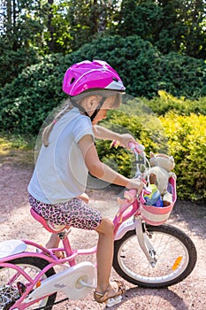 Little girl in a protective helmet rides a two wheeled pink bike in a beautiful park on a sunny summer day