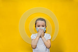 Little girl in a protective mask covers her breath with her hands on a yellow background. Protection against coronavirus