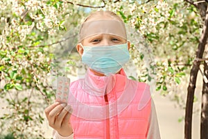 Little girl in protective mask holding pills near blooming tree. Allergy concept