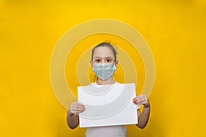 Little girl in a protective mask holds a blank sheet of paper on a yellow background. Protection against coronavirus