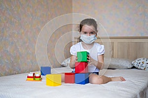 A little girl in a protective mask plays with educational toys, sitting on the bed in self-isolation, look at camera