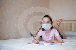 Little girl in a protective mask writes in a notebook during quarantine. Distance learning education. look at camera