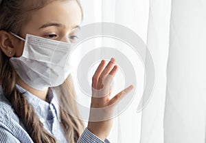 Little girl in a protective medical mask looks out the window close up