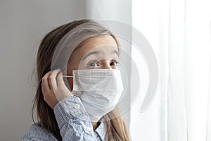 Little girl in a protective medical mask stands near the window close up