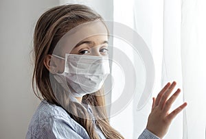 Little girl in a protective medical mask stands near the window close up