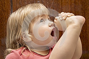 A little girl pulls her leg up to her mouth against the background of a wooden home Cabinet, stretching the muscles, childlike pla