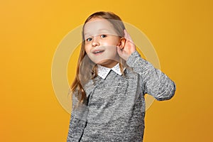 The little girl puts her hand to her ear to hear better. The child is listening to something on a yellow background