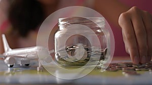 Little girl putting money into a glass jar which is on the world map with aircraft toy. Saving money for travel.