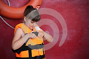 Little girl putting on orange life vest near red wall with safety ring. Space for text