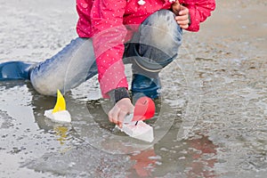 Little girl in rain boots playing with ships in the spring water puddle
