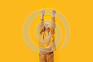 Little girl raised hands up and points index fingers up. Copy space, mock up. Vertical frame, child in yellow clothes