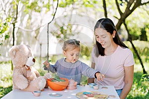 Little girl reaches for a toy cup of tea while sitting next to her mother at the table