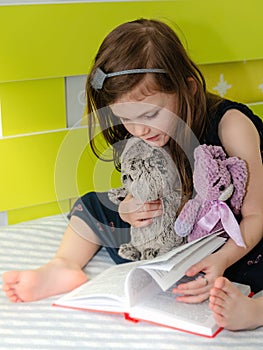 A little girl is reading a book of fairy tales to her toys in her room on the bed.