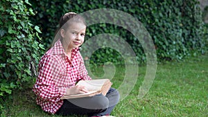 Little girl reading a book outside sitting on the green grass in the park in the meadow. Read and relax.
