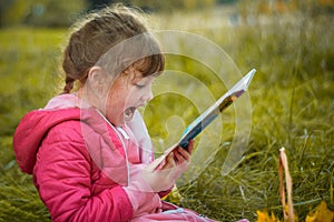 Little girl is reading a book in the park. A baby with a book is sitting on the grass. A child in a pink jacket screams gaily