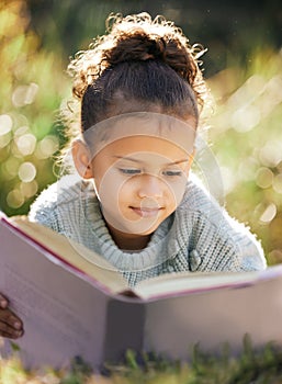 A little girl reading a book while relaxing in a park or garden. Mixed race child learning and getting an education