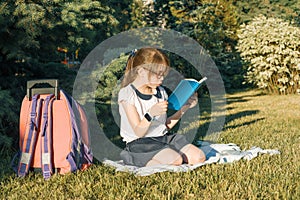 Little girl reading a book with a school backpack sitting on the grass in the park