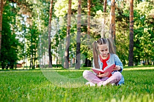 Little girl reading a book sit on grass in summer day in the outdoors