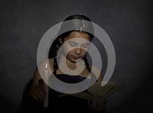 Little girl reading book and studying in candle light in dark room