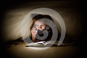 Little girl is reading a book under a blanket with a flashlight in a dark room at night